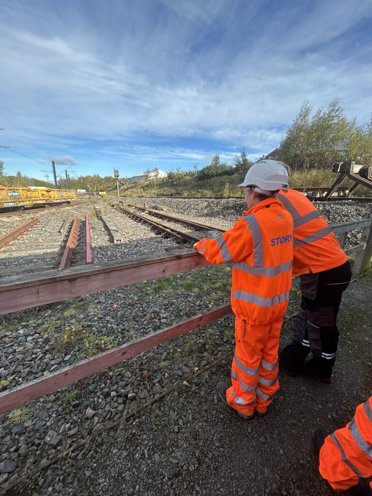 Story Contracting's employee, standing next to a young teenager, both wearing orange neon uniform of the organization, and a white helmet. They are standing in front of railway tracks