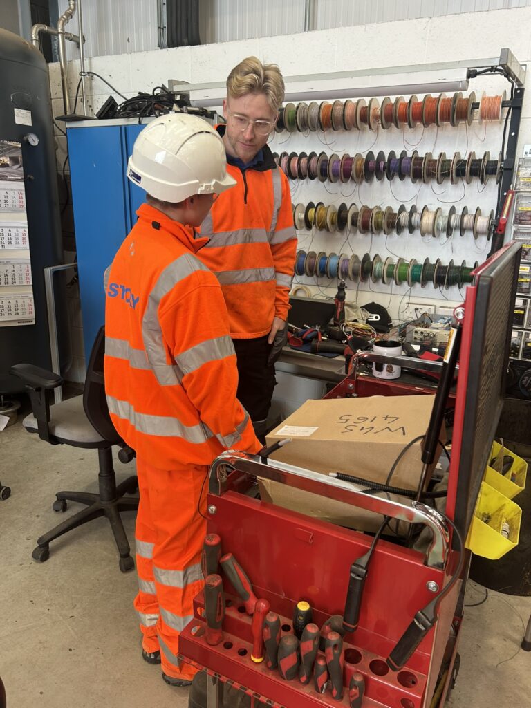An employee of Story COntracting, he has white skin and blond hair, wearing glasses, he is talking to a young teenager who wears a white helmet