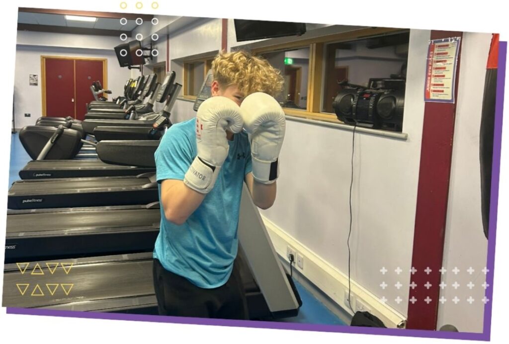 A young man wearing boxing gloves seen in a gym, surrounded by training equipment and a punching bag.